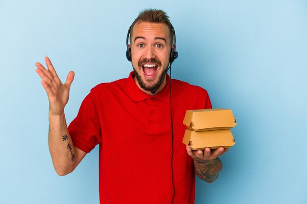 Young caucasian delivery man with tattoos holding burgers isolated on blue background  receiving a pleasant surprise, excited and raising hands.