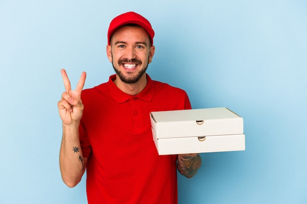 Young caucasian delivery man with tattoos holding pizzas isolated on blue background  showing number two with fingers.