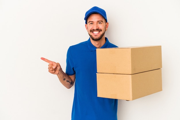 Young caucasian delivery man with tattoos isolated on white background  smiling and pointing aside, showing something at blank space.