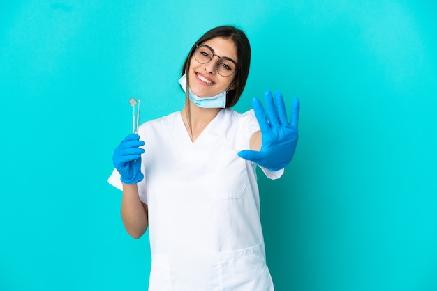 손가락으로 다섯을 세는 파란색 배경에 고립 된 도구를 들고 젊은 백인 치과 의사
