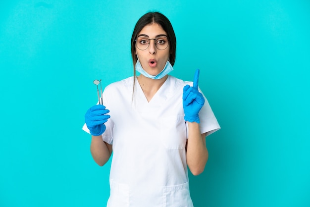 指を持ち上げながら解決策を実現することを意図して青い背景で隔離のツールを保持している若い白人歯科医の女性