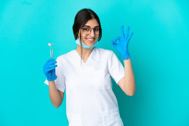 Young caucasian dentist woman holding tools isolated on blue background showing ok sign with fingers