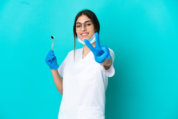 Giovane donna caucasica del dentista che tiene gli strumenti isolati su fondo blu che sorride e che mostra il segno di vittoria