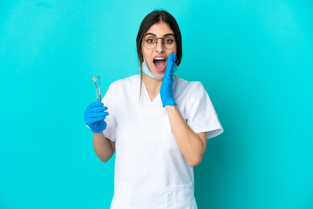 Young caucasian dentist woman holding tools isolated on blue background with surprise and shocked facial expression