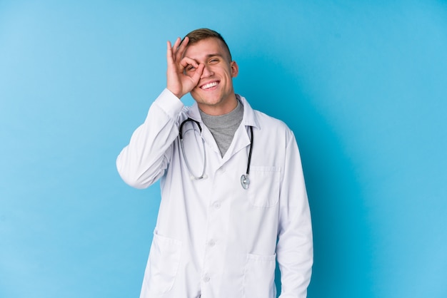 Young caucasian doctor man excited keeping ok gesture on eye.