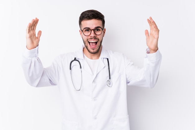 Young caucasian doctor man receiving a pleasant surprise, excited and raising hands.