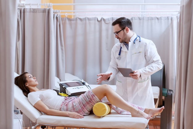 Young Caucasian doctor in white uniform holding tablet and explaining patient recovery process. Patient having leg injury and lying down on bed. Hospital interior.