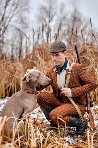 양복을 입은 젊은 백인 유럽 남자 사냥꾼은 사냥꾼을 위해 들판에 개와 함께 앉아 있습니다.