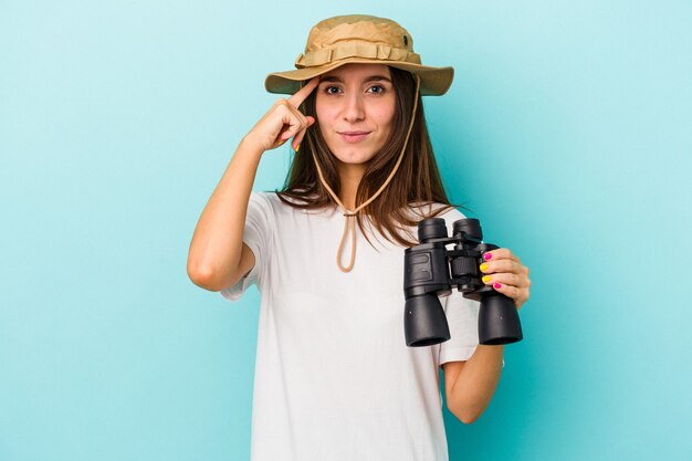 Young caucasian explorer woman holding binoculars isolated on blue background pointing temple with finger, thinking, focused on a task.