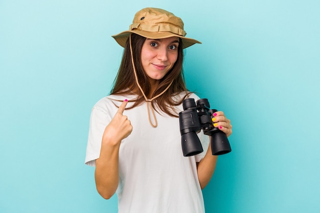 Young caucasian explorer woman holding binoculars isolated on blue background pointing with finger at you as if inviting come closer.