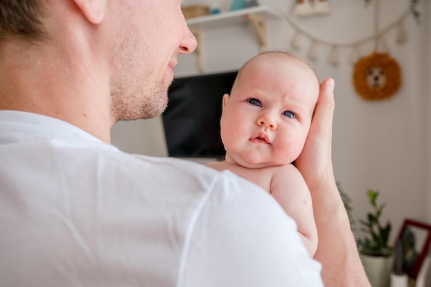 A young caucasian father gently holds a baby in a bright room at home. Happy fatherhood and the joy