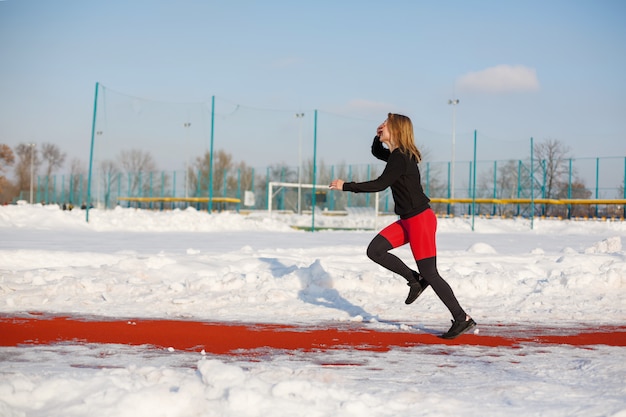 Giovane bionda femmina caucasica in ghette viola che allungano esercizio su una pista da corsa rossa in uno stadio nevoso, in forma e stile di vita sportivo