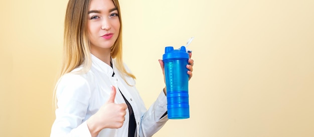 La giovane dottoressa caucasica che indossa un camice bianco tiene una bottiglia di plastica blu con acqua con il pollice in su guardando la telecamera