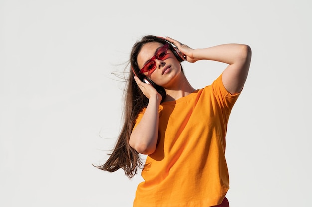 Молодая кавказка в оранжевой футболке и красных солнцезащитных очках слушает музыку в наушниках