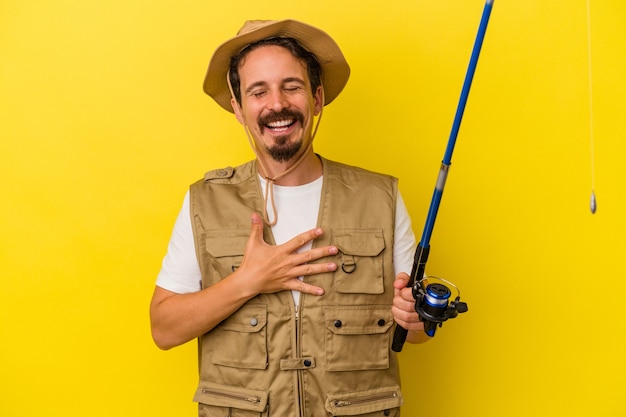 Young caucasian fisherman holding rod isolated on yellow background laughs out loudly keeping hand on chest