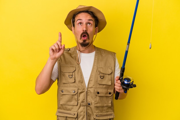 Young caucasian fisherman holding rod isolated on yellow background pointing upside with opened mouth.