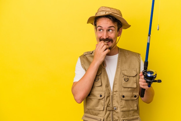 Young caucasian fisherman holding rod isolated on yellow background relaxed thinking about something looking at a copy space.