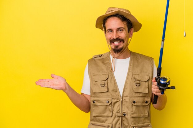 Young caucasian fisherman holding rod isolated on yellow background showing a copy space on a palm and holding another hand on waist