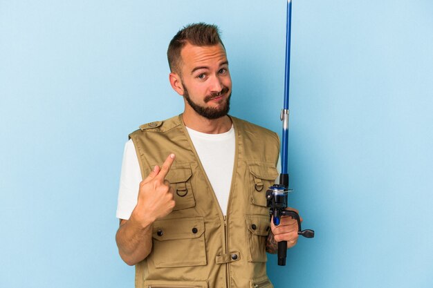 Young caucasian fisherman with tattoos holding rod isolated on blue background pointing with finger at you as if inviting come closer