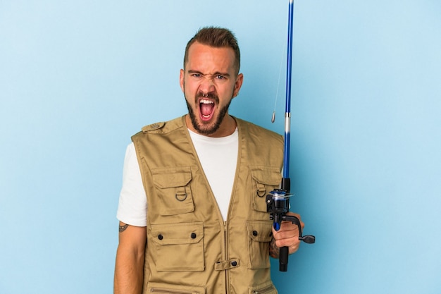 Young caucasian fisherman with tattoos holding rod isolated on blue background  screaming very angry and aggressive.