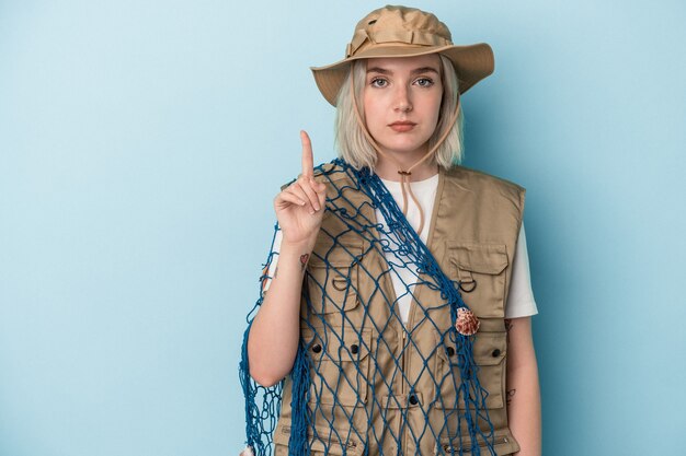 Young caucasian fisherwoman holding a net isolated on blue background showing number one with finger.