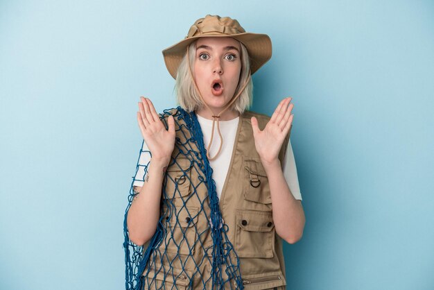 Young caucasian fisherwoman holding a net isolated on blue background surprised and shocked.