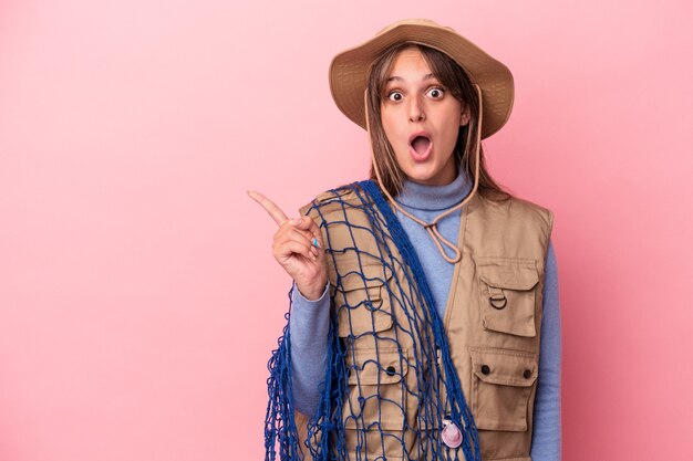 Young caucasian fisherwoman holding a net isolated on pink background pointing to the side