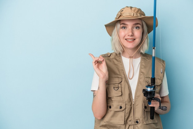 Young caucasian fisherwoman holding a rod isolated on blue background smiling and pointing aside, showing something at blank space.