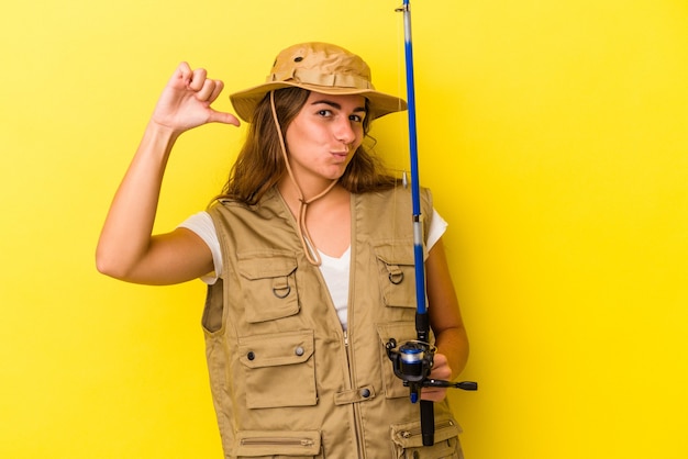 Young caucasian fisherwoman holding a rod isolated on yellow background  feels proud and self confident, example to follow.