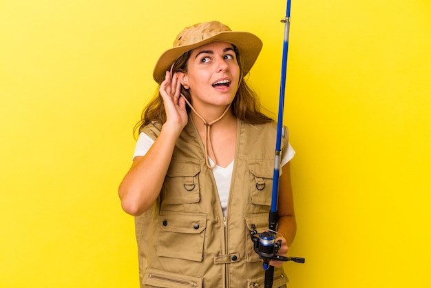 Photo young caucasian fisherwoman holding a rod isolated on yellow background  trying to listening a gossip.