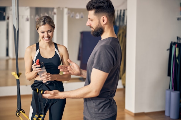 Young Caucasian fitness instructor demonstrating the TRX straps to a smiling woman at a gym