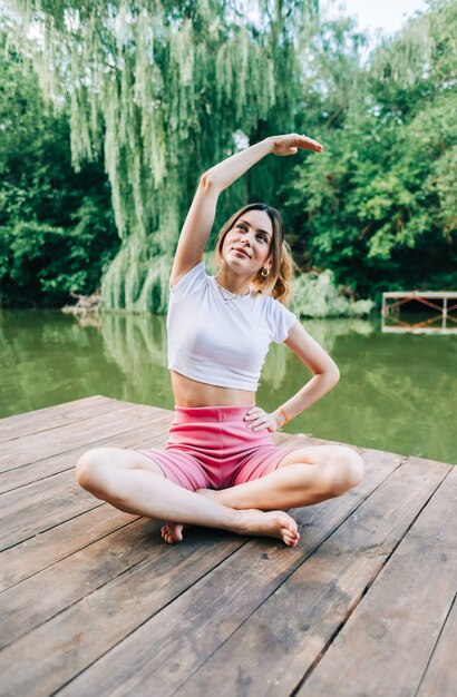 Фото Молодая кавказская женщина фитнеса делает йогу, медитирует на открытом воздухе, на озере деревянной пристани в летний день.