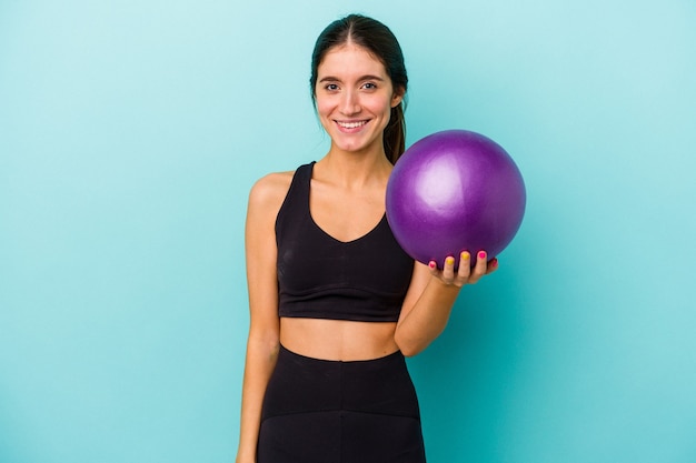 Foto giovane donna caucasica fitness in possesso di una palla isolata su sfondo blu felice, sorridente e allegra.