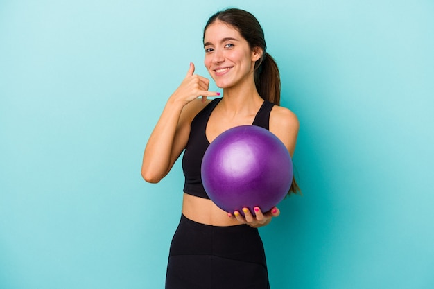 Young caucasian fitness woman holding a ball isolated on blue background showing a mobile phone call gesture with fingers.