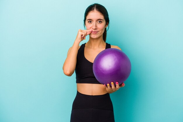 Young caucasian fitness woman holding a ball isolated on blue background with fingers on lips keeping a secret.