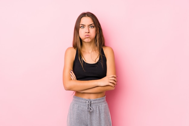 Young caucasian fitness woman posing in a pink wall blows cheeks, has tired expression. Facial expression concept.