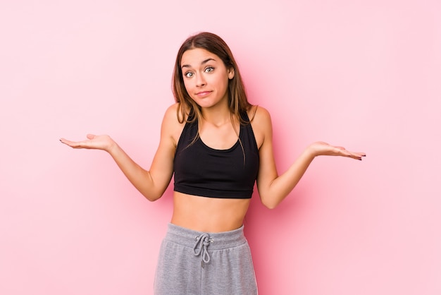 Молодая кавказская женщина фитнеса представляя в розовой стене сомневаясь и пожимая плечами в спрашивая жесте.