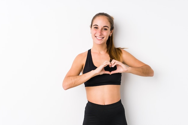 Young caucasian fitness woman posing in a white background smiling and showing a heart shape with hands.