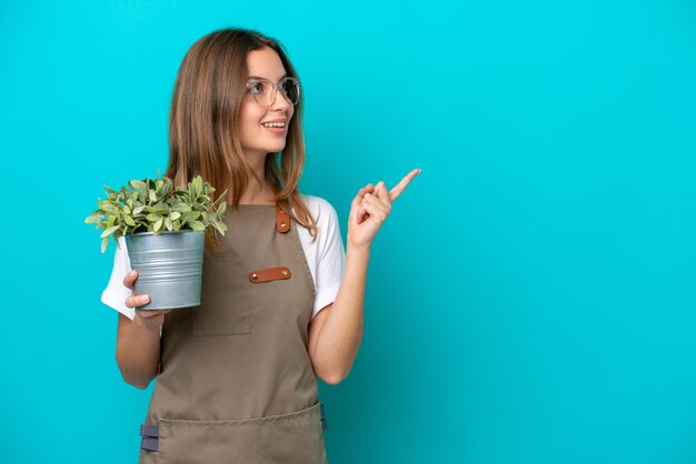 파란색 배경에 격리된 식물을 들고 있는 백인 정원사 여성은 손가락을 들어올리면서 해결책을 실현하고자 합니다.