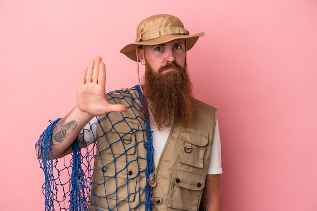 Foto giovane pescatore caucasico dello zenzero con la barba lunga che tiene una rete isolata su fondo rosa