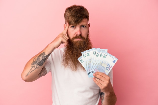 Young caucasian ginger man with long beard holding banknotes isolated on pink background pointing temple with finger, thinking, focused on a task.