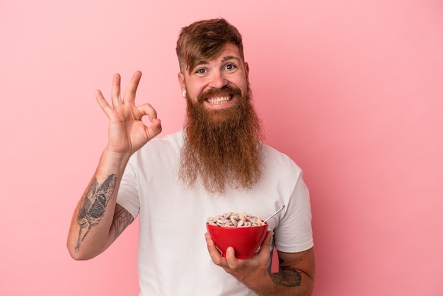Young caucasian ginger man with long beard holding a bowl of cereales isolated on pink background cheerful and confident showing ok gesture.
