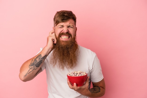 Young caucasian ginger man with long beard holding a bowl of cereales isolated on pink background covering ears with hands.