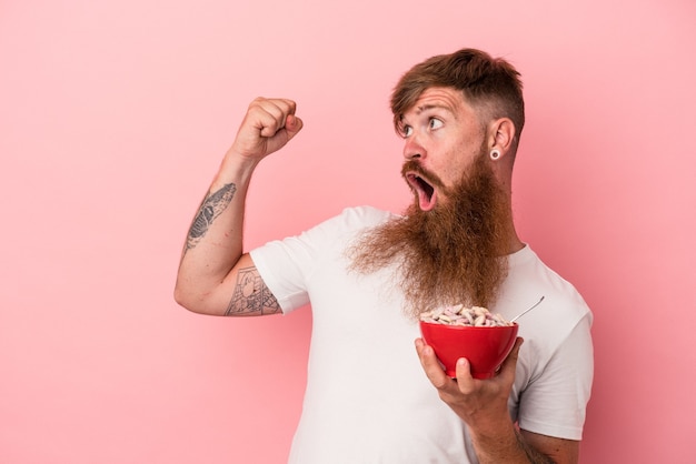 Young caucasian ginger man with long beard holding a bowl of cereales isolated on pink background raising fist after a victory, winner concept.