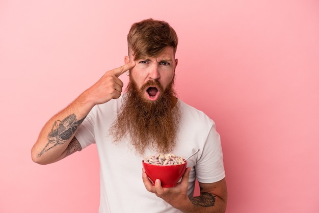 Young caucasian ginger man with long beard holding a bowl of cereales isolated on pink background showing a disappointment gesture with forefinger.