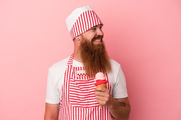 Молодой кавказский рыжий мужчина с длинной бородой, держащий мороженое на розовом фоне, смотрит в сторону улыбающимся, веселым и приятным.