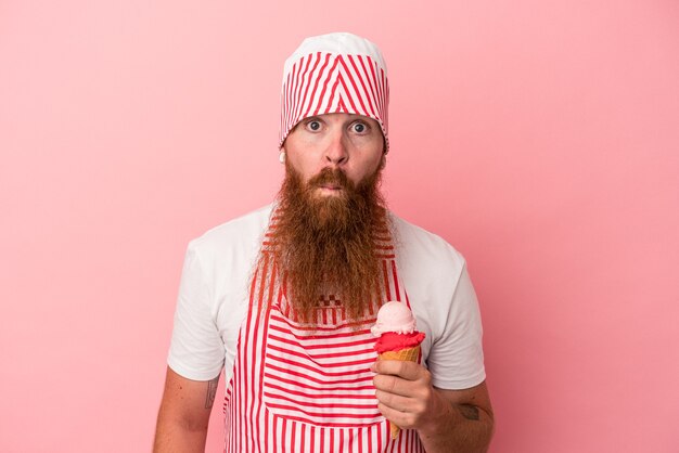 Young caucasian ginger man with long beard holding an ice cream isolated on pink background shrugs shoulders and open eyes confused.