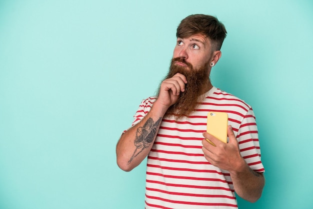 Giovane uomo caucasico dello zenzero con la barba lunga che tiene un telefono cellulare isolato su priorità bassa blu