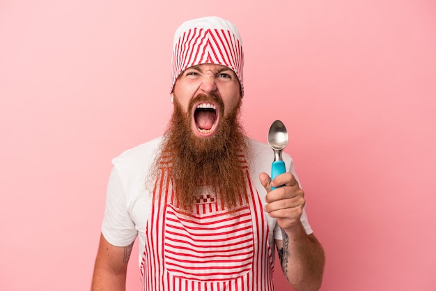 Young caucasian ginger man with long beard holding a scoop isolated on pink background screaming very angry and aggressive.