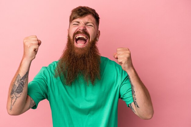 Young caucasian ginger man with long beard isolated on pink background cheering carefree and excited. Victory concept.